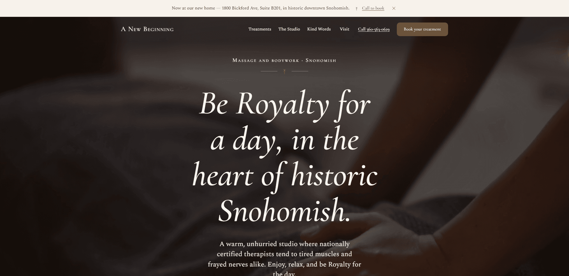 Homepage of A New Beginning: a cream announcement bar reading 'Now at our new home - 1800 Bickford Ave, Suite B201, in historic downtown Snohomish.' with a 'Call to book' link and a close button, above a dark, soft-focus still of a hand resting on a grey blanket, under a top bar carrying the small-capitals serif 'A New Beginning' wordmark, navigation reading Treatments, The Studio, Kind Words, and Visit, a 'Call' phone number link, and a brown 'Book your treatment' button, above the centered small-capitals kicker 'Massage and bodywork - Snohomish' over a fine ornamental divider, the oversized italic serif headline 'Be Royalty for a day, in the heart of historic Snohomish.', and a paragraph about a warm, unhurried studio where nationally certified therapists tend to tired muscles and frayed nerves, running past the bottom edge.