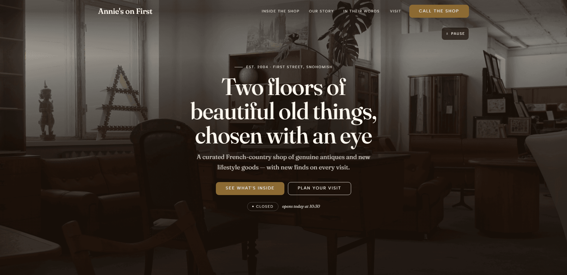 Homepage of Annie's on First: a dark, warm wash over a video still of an antique showroom of wooden cabinets, leather chairs, framed prints, a large monstera plant, and a decorative letter A by a bright window, under a top bar carrying the serif 'Annie's on First' wordmark, navigation reading Inside the shop, Our story, In their words, and Visit, and a bronze 'Call the shop' button, above the centered kicker 'Est. 2004 - First Street, Snohomish' on a short rule, the large serif headline 'Two floors of beautiful old things, chosen with an eye', a line about a curated French-country shop of genuine antiques and new lifestyle goods, a bronze 'See what's inside' button beside an outlined 'Plan your visit' button, a 'Closed' status pill beside the italic line 'opens today at 10:30', and a pause control for the background video in the corner.