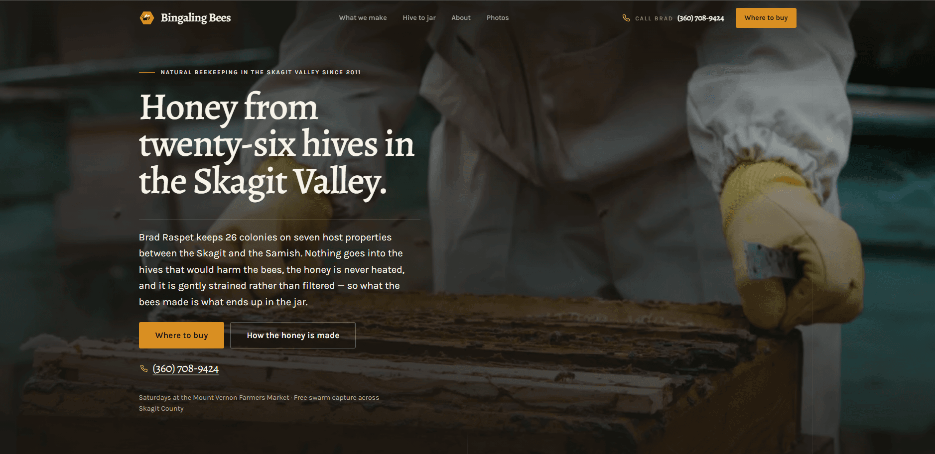 Homepage of Bingaling Bees: a dark, warm-toned page over a photograph of a beekeeper in a white suit and yellow gloves working a hive frame with a hive tool, smoke drifting across it, under a top bar carrying a hexagonal bee mark, navigation reading What we make, Hive to jar, About, and Photos, a 'Call Brad' phone number, and a 'Where to buy' button, above the kicker 'Natural beekeeping in the Skagit Valley since 2011', the serif headline 'Honey from twenty-six hives in the Skagit Valley.', a paragraph about 26 colonies kept on seven host properties and honey that is never heated, and 'Where to buy' and 'How the honey is made' actions beside a note about Saturdays at the Mount Vernon Farmers Market.