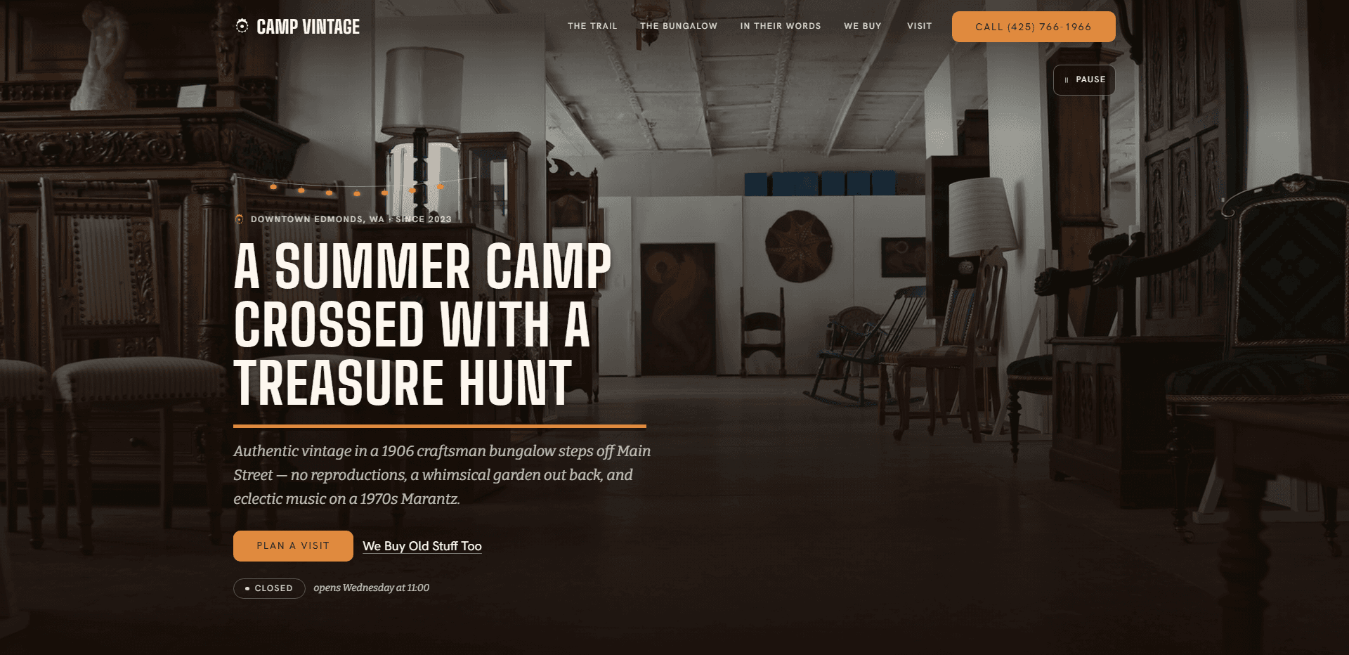 Homepage of Camp Vintage: a dark wash over a video still of a white-walled antique showroom of carved dark wood cabinets, chairs, a rocking chair, tall lamps, and a round wall hanging beneath a string of small lights, under a top bar carrying a small round mark beside the condensed capitals 'Camp Vintage' wordmark, navigation reading The trail, The bungalow, In their words, We buy, and Visit, an orange 'Call' phone number button, and a pause control for the background video beneath it, above the small capitals kicker 'Downtown Edmonds, WA - Since 2023', the oversized condensed capitals headline 'A summer camp crossed with a treasure hunt' over an orange rule, an italic line about authentic vintage in a 1906 craftsman bungalow steps off Main Street, with no reproductions, a whimsical garden out back, and eclectic music on a 1970s Marantz, an orange 'Plan a visit' button beside an underlined 'We Buy Old Stuff Too' link, and a 'Closed' status pill beside the italic line 'opens Wednesday at 11:00'.