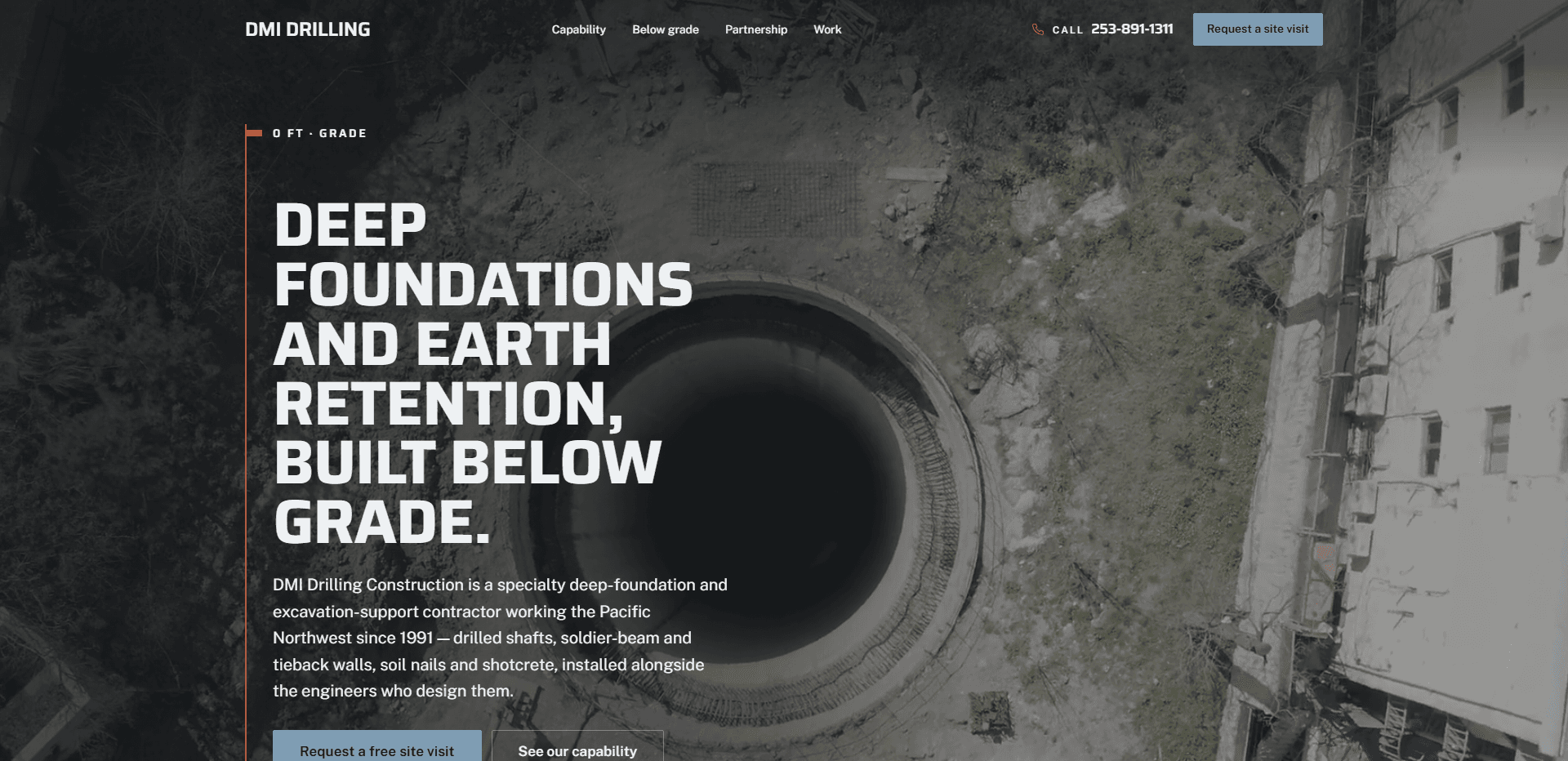 Homepage of DMI Drilling Construction: a dark wash over an aerial view looking straight down into a wide, round drilled shaft ringed with rebar on a dirt and gravel site beside a white multi-story building, under a top bar carrying the bold 'DMI Drilling' wordmark, navigation reading Capability, Below grade, Partnership, and Work, a 'Call' phone number, and a steel-blue 'Request a site visit' button, above the kicker '0 ft - Grade' at the top of an orange depth rule running down the left side, the oversized heavy capitals headline 'Deep foundations and earth retention, built below grade.', a paragraph about a specialty deep-foundation and excavation-support contractor working the Pacific Northwest since 1991 on drilled shafts, soldier-beam and tieback walls, and soil nails and shotcrete, and a steel-blue 'Request a free site visit' button beside an outlined 'See our capability' button, both cut off at the bottom edge.