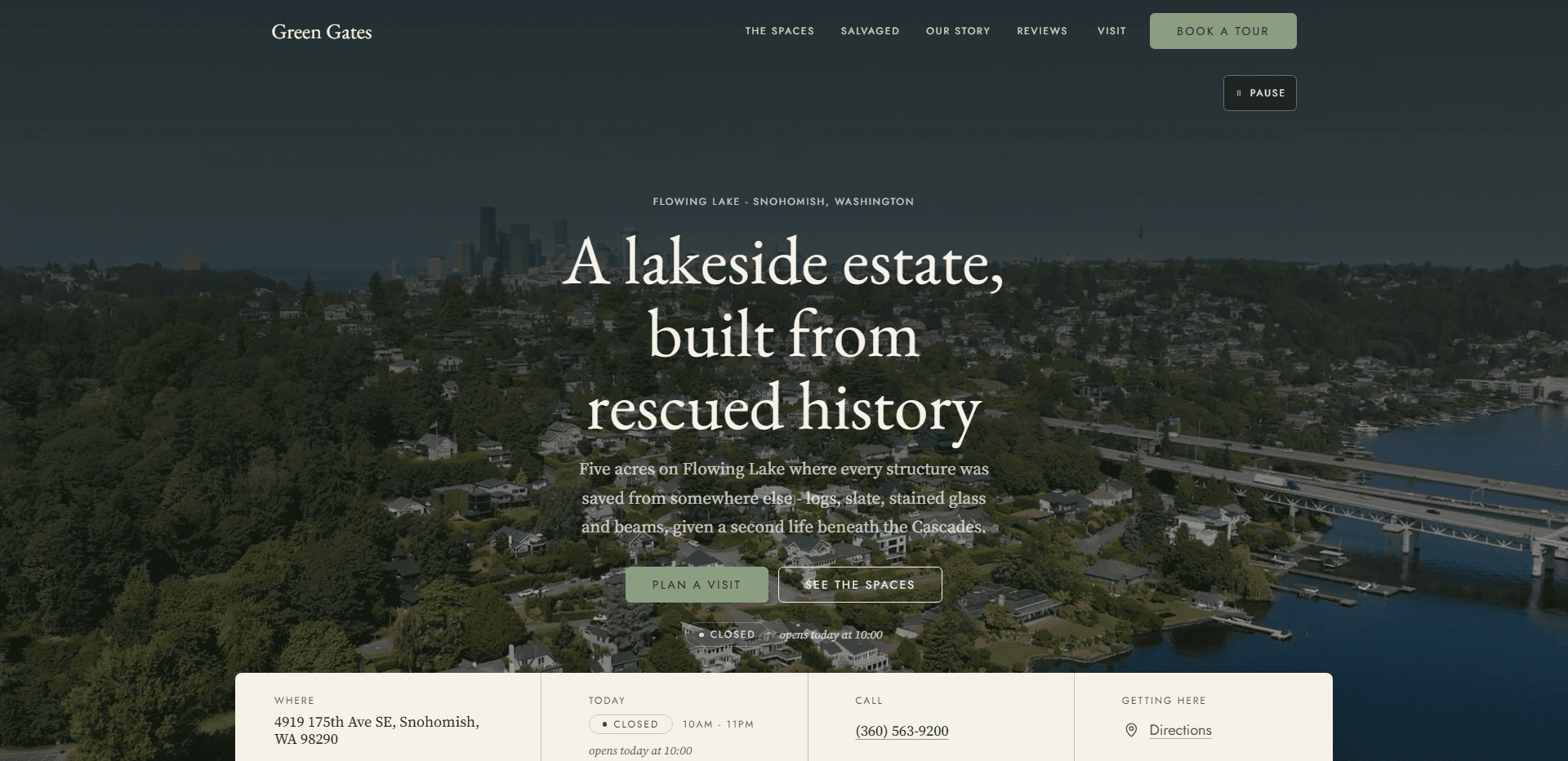 Homepage of Green Gates: a dark wash over an aerial video still of a wooded lakeside neighborhood with docks along the shore, a highway bridge crossing the water, and a city skyline on the horizon, under a top bar carrying the serif 'Green Gates' wordmark, navigation reading The Spaces, Salvaged, Our Story, Reviews, and Visit, and a sage green 'Book a tour' button, above a 'Pause' control for the background video, the centered kicker 'Flowing Lake - Snohomish, Washington', the large serif headline 'A lakeside estate, built from rescued history', a line about five acres on Flowing Lake where every structure, from logs and slate to stained glass and beams, was saved from somewhere else and given a second life beneath the Cascades, a sage green 'Plan a visit' button beside an outlined 'See the spaces' button, a status line reading Closed and opens today at 10:00, and a cream information bar listing the address, today's hours, the phone number, and a directions link.