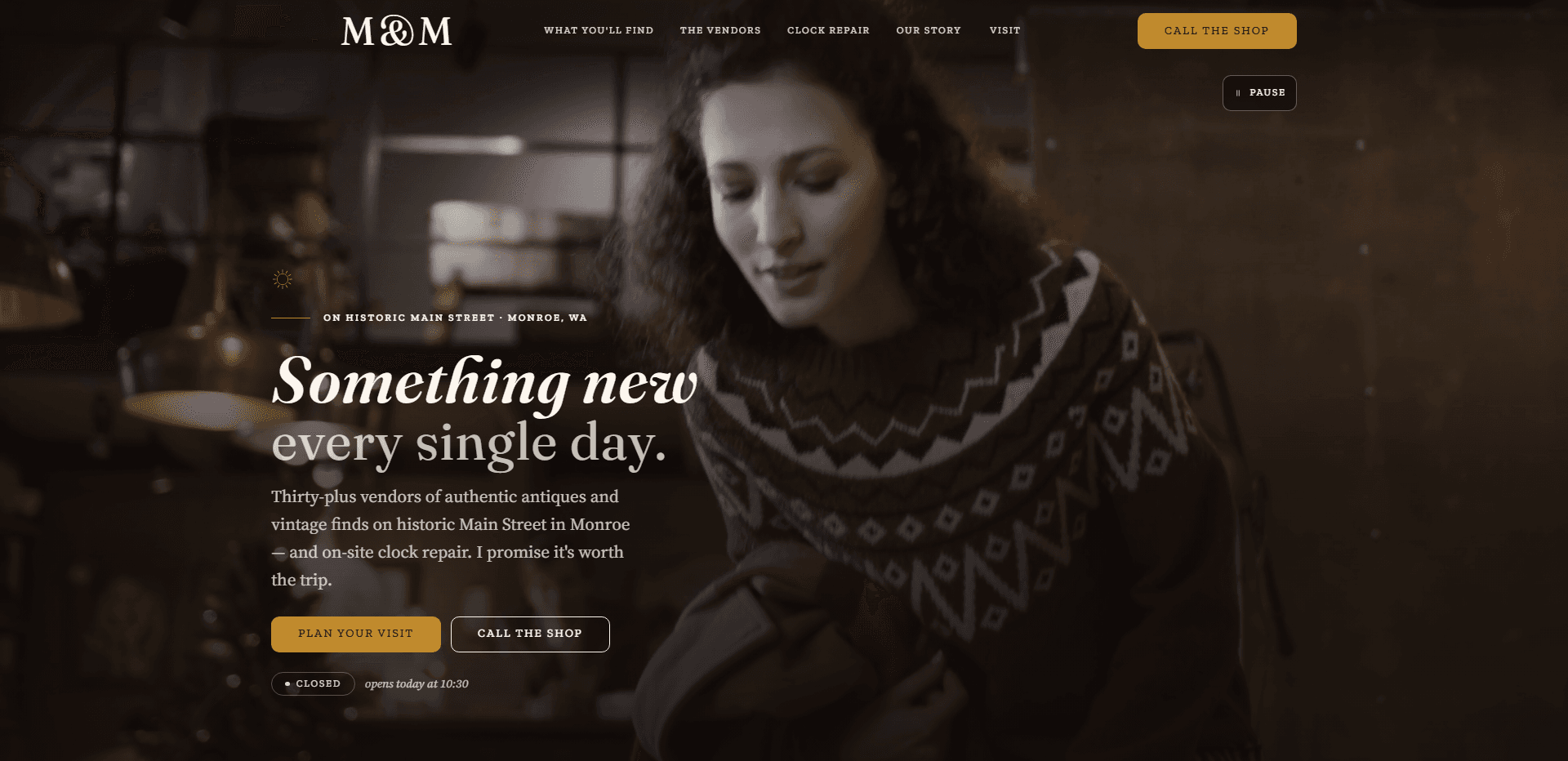 Homepage of M & M Antiques & Collectibles: a dark, warm video still of a curly-haired woman in a patterned red-and-white wool sweater looking down at something in her hands inside a dimly lit shop with a hanging brass lamp, under a top bar carrying the serif 'M&M' wordmark, navigation reading What you'll find, The vendors, Clock repair, Our story, and Visit, and a gold 'Call the shop' button, above a small sun mark and the kicker 'On historic Main Street - Monroe, WA' on a gold rule, the large serif headline 'Something new every single day.' with its first line in italic, a paragraph about thirty-plus vendors of antiques and vintage finds on historic Main Street in Monroe and on-site clock repair, a gold 'Plan your visit' button beside an outlined 'Call the shop' button, a 'Closed' status pill beside the italic line 'opens today at 10:30', and a pause control for the background video in the corner.