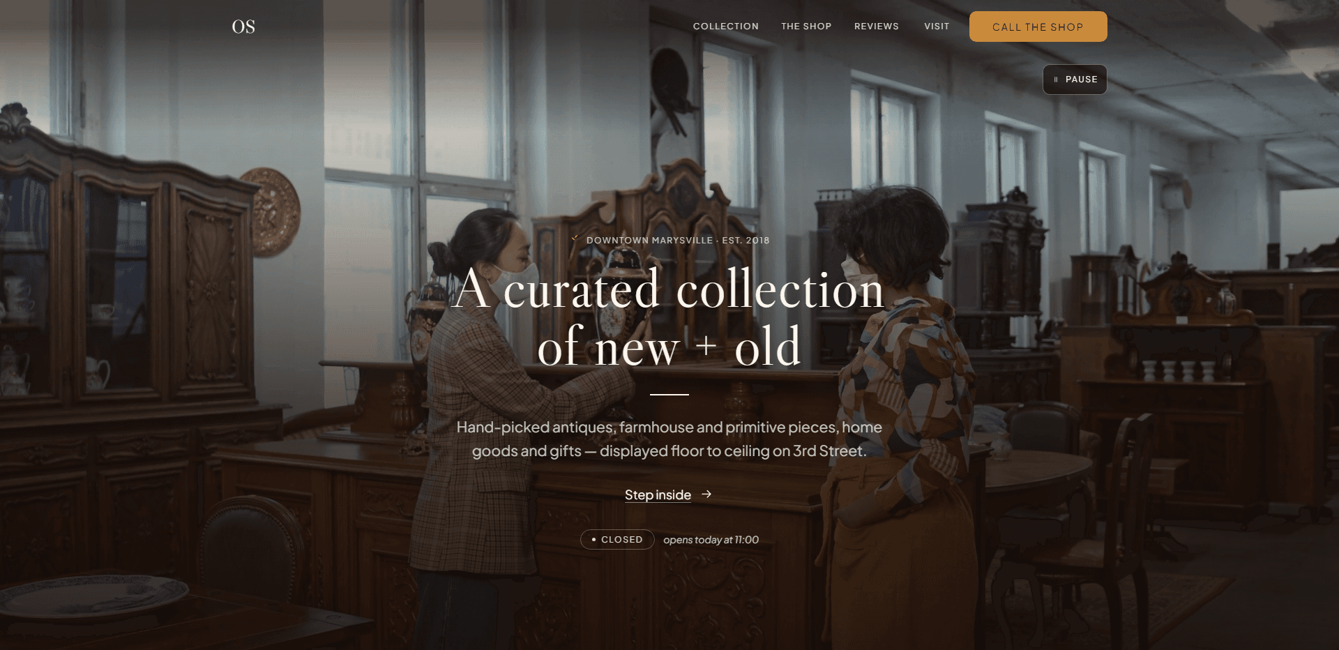 Homepage of Old Soul: a dark wash over a video still of two women in face masks examining a black-and-gold lidded urn among carved antique cabinets and sideboards in a tall-windowed warehouse, under a top bar carrying the serif 'OS' monogram, navigation reading Collection, The shop, Reviews, and Visit, and an amber 'Call the shop' button, above the centered kicker 'Downtown Marysville - Est. 2018', the large light serif headline 'A curated collection of new + old' over a short white rule, a line about hand-picked antiques, farmhouse and primitive pieces, home goods, and gifts displayed floor to ceiling on 3rd Street, a 'Step inside' link with an arrow, a 'Closed' status pill beside the italic line 'opens today at 11:00', and a pause control for the background video in the corner.