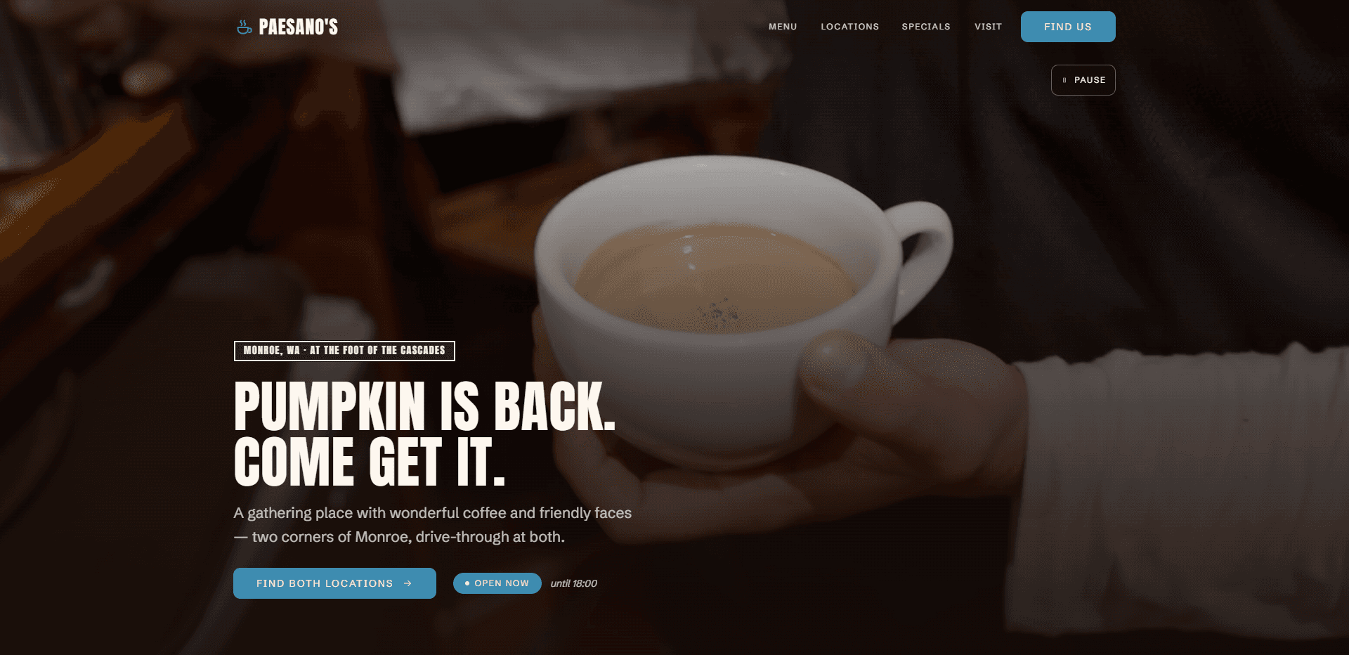 Homepage of Paesano's Coffee: a near-black wash over a soft-focus video still of a hand holding a white cup of latte, under a top bar carrying a small coffee cup mark beside the heavy condensed capitals 'Paesano's' wordmark, navigation reading Menu, Locations, Specials, and Visit, and a blue 'Find us' button, with a pause control for the background video beneath it, above a boxed kicker 'Monroe, WA - At the foot of the Cascades', the oversized condensed capitals headline 'Pumpkin is back. Come get it.', a line about a gathering place with wonderful coffee and friendly faces at two corners of Monroe, drive-through at both, a blue 'Find both locations' button, and a blue 'Open now' status pill beside an italic closing time.
